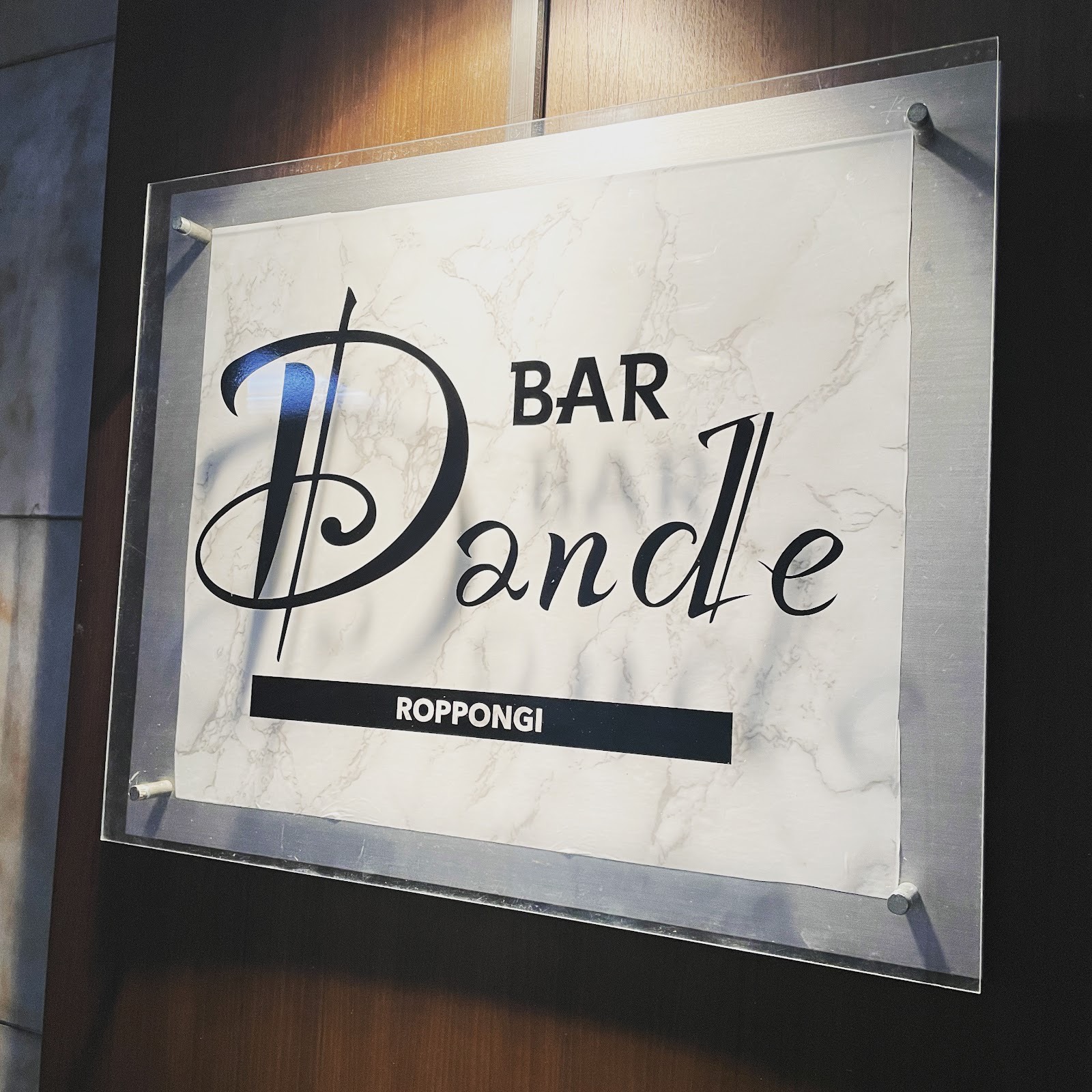 「BAR Dande ROPPONGI」の看板。木目の壁に透明アクリル板で留めた白い大理石調のプレートに、筆記体の黒い「Dande」、その上に「BAR」、下に黒帯の「ROPPONGI」。