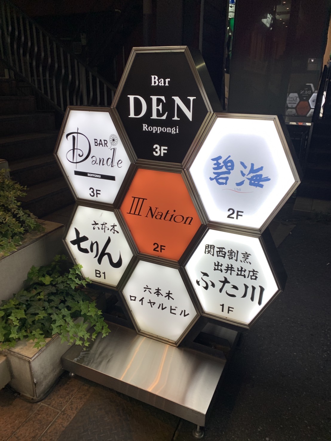 夜の六本木ロイヤルビル前に立つ六角形の館内案内看板。左上に「BAR Dande 3F」のパネル。